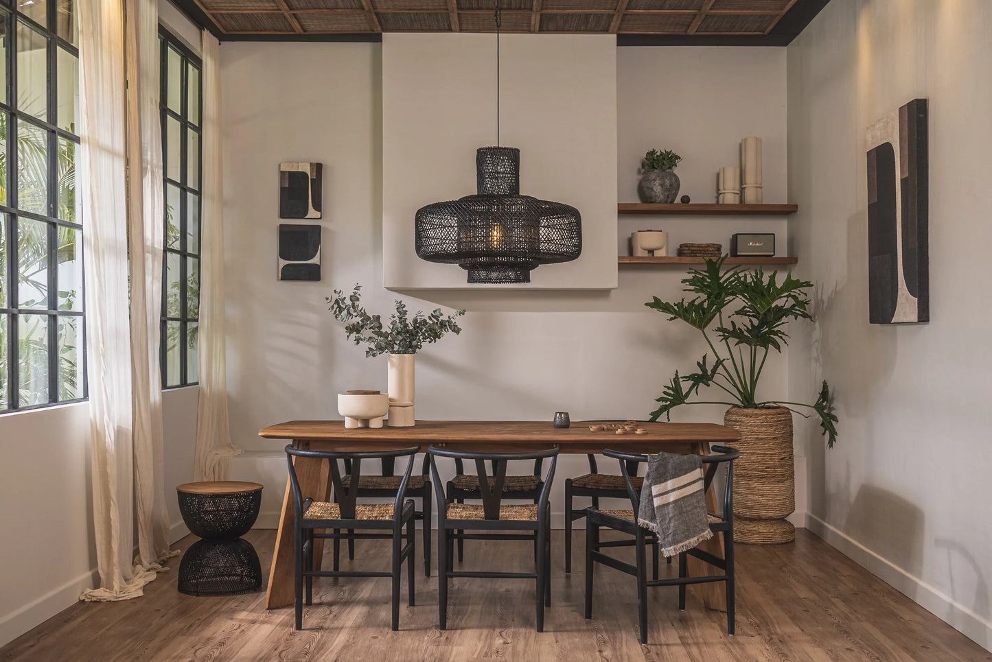 Table de cuisine en bois massif de teck recyclé, modèle Grace de la collection CRUSOË X dBodhi. Entourée de chaises de type "wishbone" noires dans une salle à manger d'inspiration mid-century tropical.